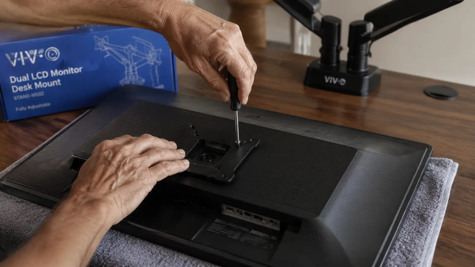 VESA mounting plate being attached to the back of a monitor with four screws, desk arm visible in the background