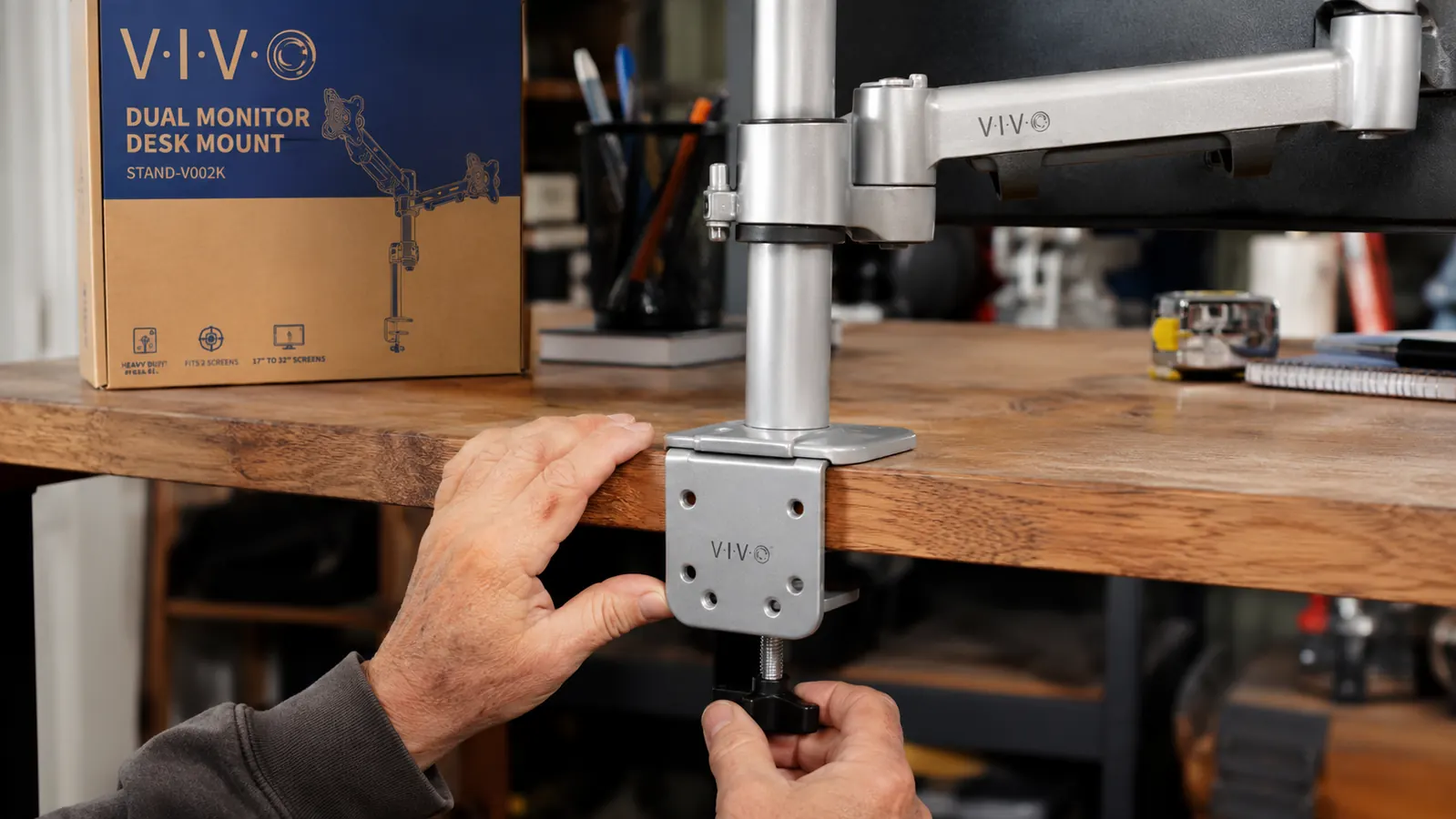 Close-up of the VIVO STAND-V002 dual monitor arm C-clamp clamped to the edge of a solid wood desk, showing the steel construction and adjustment knobs